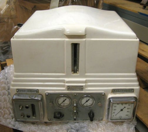 Figure 1 — The front of a modern ESCO Porti-Boy: a clear graduated glass tank above a stainless base, with the pressure gauge and the separate pressure and rate-of-flow control knobs on the panel. The two dia…