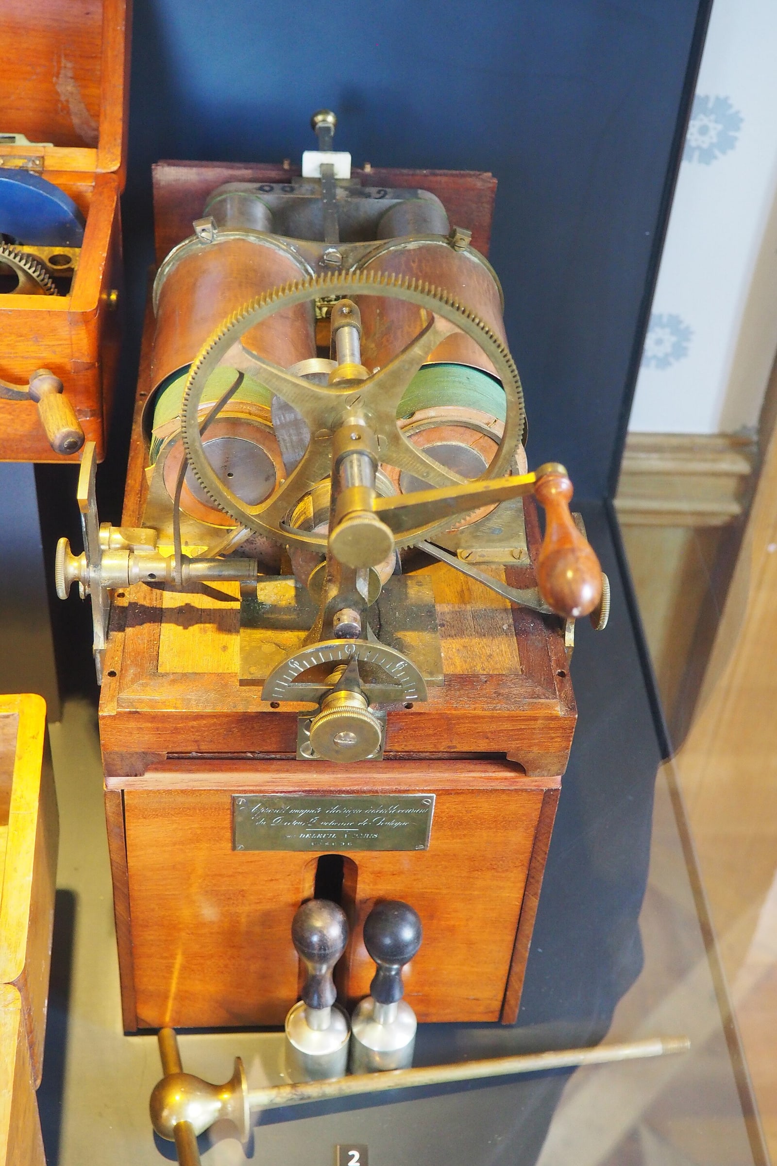 Figure 2 — A cased magneto-electric machine as it sits on a museum shelf, brass gearing and crank uppermost, the electrode handles and their cords laid in front. For identification, work from the top down: ma…